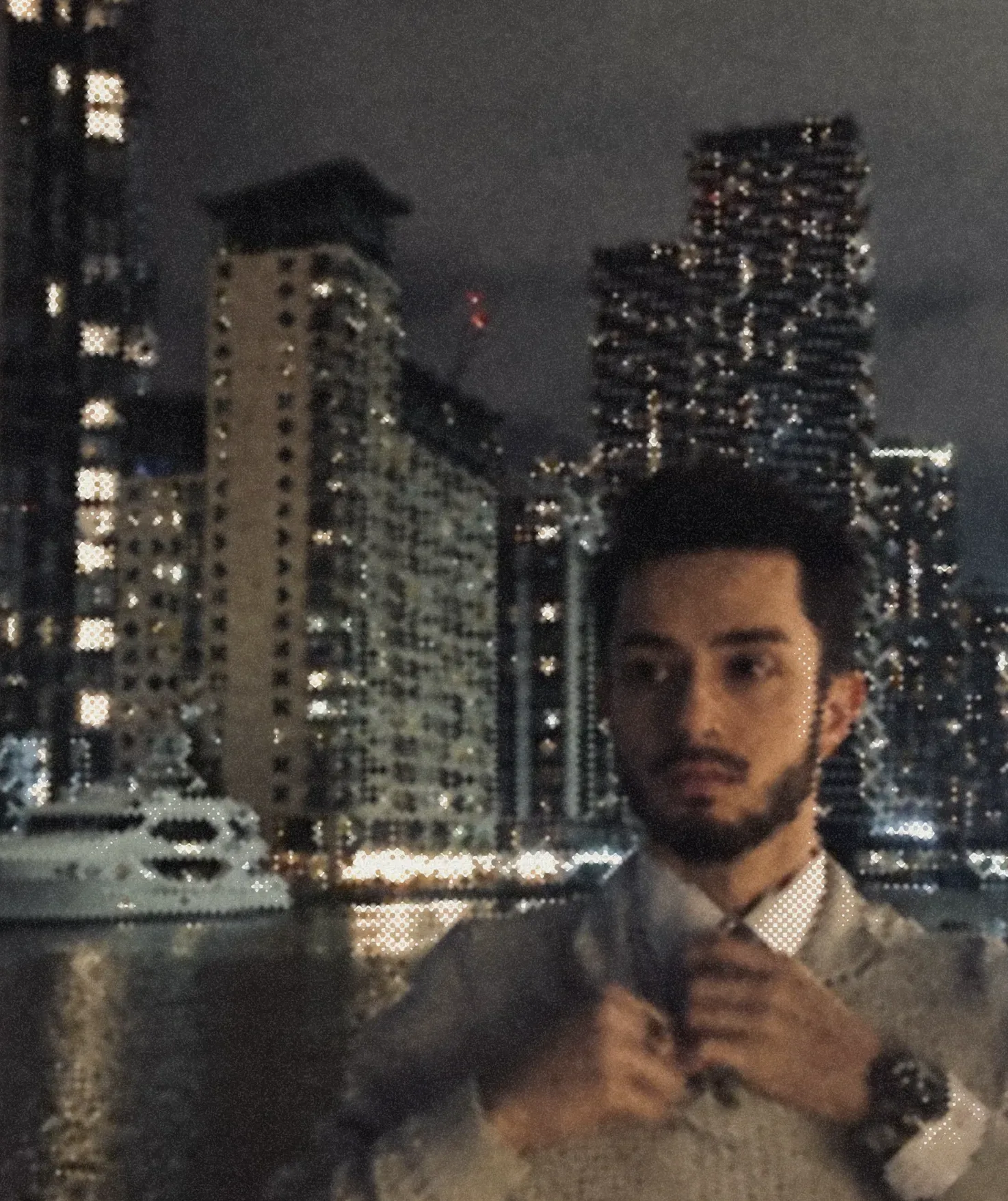 A man doing up his tie at a window, a city of lit towers across the water behind him.