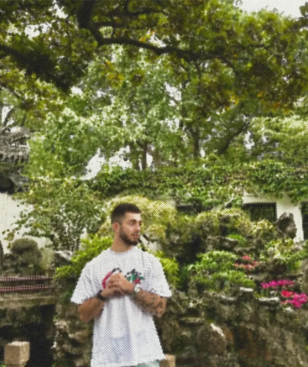 A man standing in a garden under a canopy of trees, stone and flowers behind him.